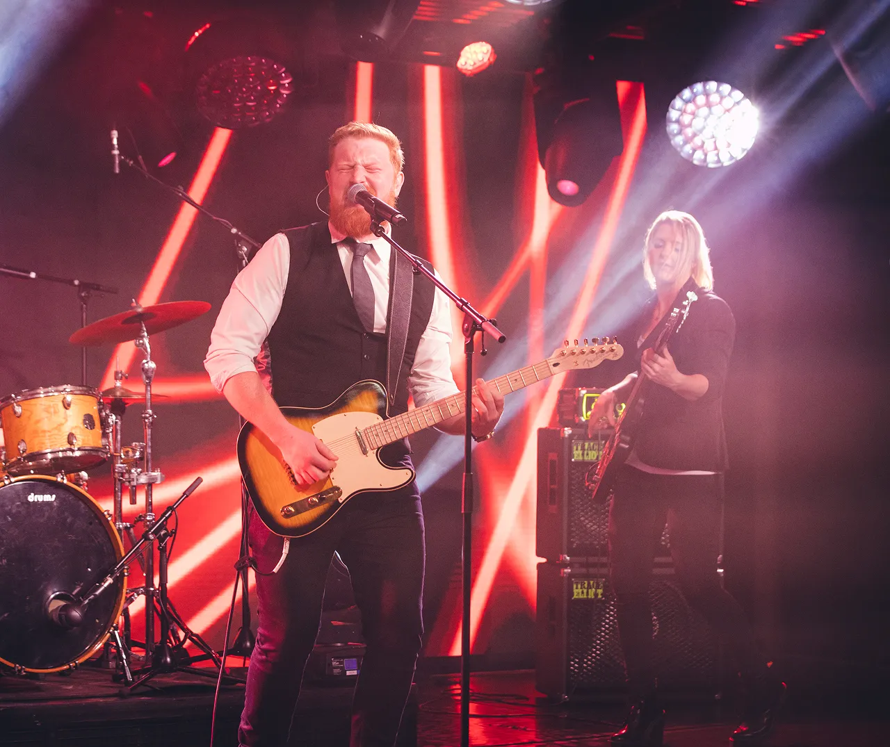 Male singer with beard wearing a vest and tie playing an electric guitar and female bassist performing onstage with red and white stage lights.